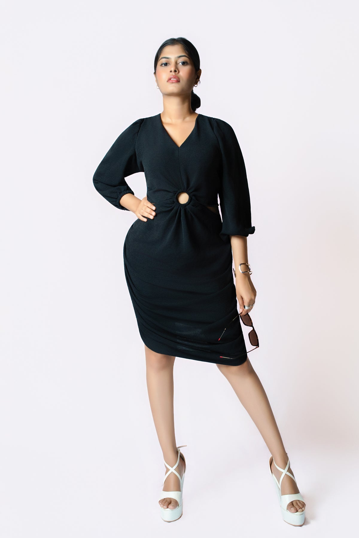 Noir Ring Detail Dress: black V-neck knee-length dress with puff sleeves and a gold ring cut-out at the waist, front view, hand on hip